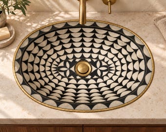 Op de afbeelding: Een ovale wastafel met een herhalend zwart-wit geometrisch waaierpatroon aan de binnenzijde. De waskom is afgewerkt met een metallic gouden rand en heeft in het midden een bijpassende ronde gouden afvoerplug.