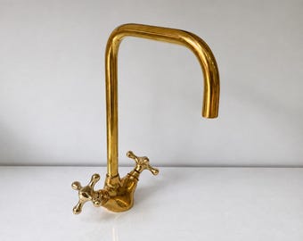 May include: A polished brass kitchen faucet featuring a high-arched spout and two cross-shaped handles. The metal finish is reflective and gold-toned, with a curved neck design and a central base that connects the spout to the two side handles.