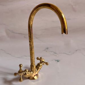 Può includere: Un rubinetto da cucina dorato con un becco ad arco alto e due maniglie a forma di croce.
