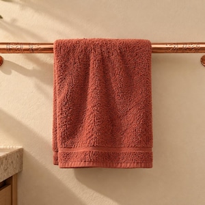 May include: A copper-toned metal towel bar with engraved leaf patterns on the ends, mounted to the wall. A solid terracotta-colored cotton bath towel is draped over the center of the bar, hanging down in a straight, folded position.