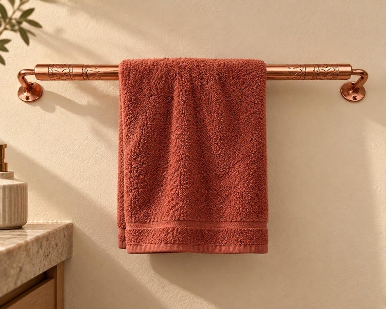 May include: A copper-toned metal towel bar with engraved leaf patterns on the ends, mounted to the wall. A solid terracotta-colored cotton bath towel is draped over the center of the bar, hanging down in a straight, folded position.