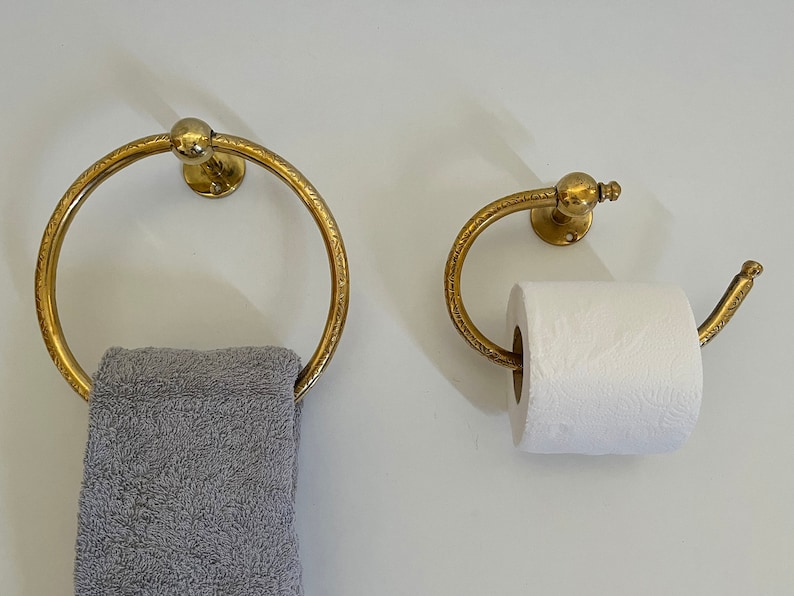 May include: A matching set of antique brass bathroom fixtures. The set includes a towel ring with a gray towel and a toilet paper holder with a roll of white toilet paper. Both fixtures are mounted on a white wall.