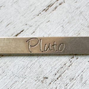May include: A silver-colored bar necklace with the name "Pluto" engraved in a cursive font. The bar is suspended from a delicate chain. The necklace is displayed on a white, textured surface.