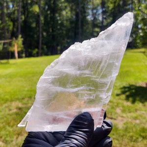 Puede incluir: Un gran cristal transparente y translúcido con un borde afilado y puntiagudo. El cristal tiene una textura en capas y estriada y se sostiene en una mano enguantada.