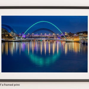 May include: A night view of a bridge with a large arch illuminated in green and purple lights. The bridge is reflected in the water below, creating a symmetrical pattern of lights. The city skyline is visible in the background.