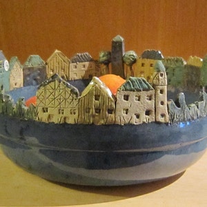 Può includere: Una ciotola in ceramica blu con una scena di una piccola città con case e alberi. La ciotola è decorata con una glassa blu e ha un bordo bianco.