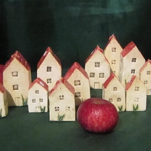 Può includere: Una collezione di piccole case in ceramica color crema con tetti rossi e piccole finestre. Le case sono disposte con mele rosse su una superficie verde, creando una scena di villaggio in miniatura. Alcune case hanno erba verde dipinta.