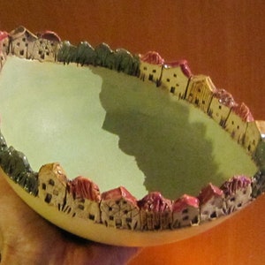 Puede incluir: Un cuenco de cerámica con un esmalte verde y un borde decorado con pequeñas casas y árboles. Las casas están pintadas en tonos de rojo, rosa, amarillo y marrón.