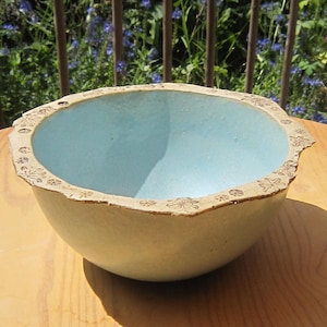 Può includere: Una ciotola in ceramica blu chiaro con un bordo bianco e un motivo floreale decorativo. La ciotola è appoggiata su una superficie di legno.