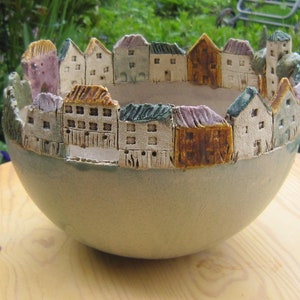 May include: A ceramic bowl with a unique design featuring a miniature village around the rim. The bowl is light gray with a textured surface and the houses are painted in various colors.