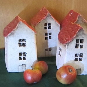 May include: Four ceramic house figurines with red roofs and square windows. The houses are white with a textured finish. Three red and yellow apples are in front of the houses, resting on a green surface.