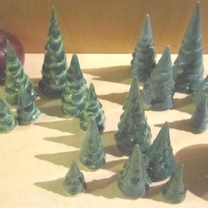 Puede incluir: Una colección de pequeñas figuras de cerámica en forma de cono en varios tonos de verde oscuro, dispuestas en grupo. Las figuras se asemejan a árboles de hoja perenne estilizados con superficies texturizadas. Dos manzanas grandes, redondas y de color rojo oscuro se colocan a los lados.