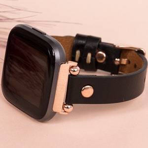 May include: A smartwatch with a dark gray square screen and a black leather strap. The watch features rose gold metal hardware, including the connector lugs, a circular stud on the strap, and a metal buckle closure for the wristband.