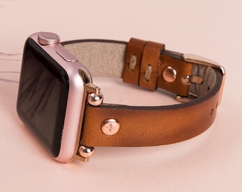 May include: A smartwatch with a rose gold metallic case and a black screen, attached to a brown leather strap. The strap features rose gold-toned metal rivets, a matching buckle, and two leather loops to secure the end of the band.