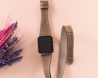 May include: A smartwatch with a rectangular black screen and a metallic silver casing, featuring a long, tan leather wrap-around watch band with white stitching and a silver buckle, positioned next to a small bouquet of dried pink and purple wheat.
