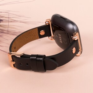 May include: A black smartwatch with a smooth leather strap and rose gold-toned metal hardware. The watch features a dark, rounded rectangular case with visible sensors on the back and a buckle closure on the strap, showcasing a refined design.
