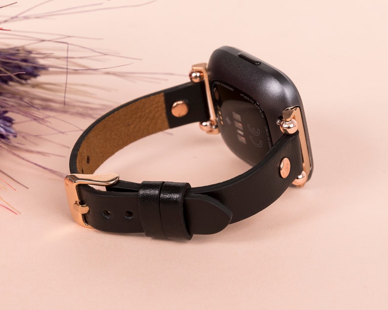 May include: A black smartwatch with a smooth leather strap and rose gold-toned metal hardware. The watch features a dark, rounded rectangular case with visible sensors on the back and a buckle closure on the strap, showcasing a refined design.