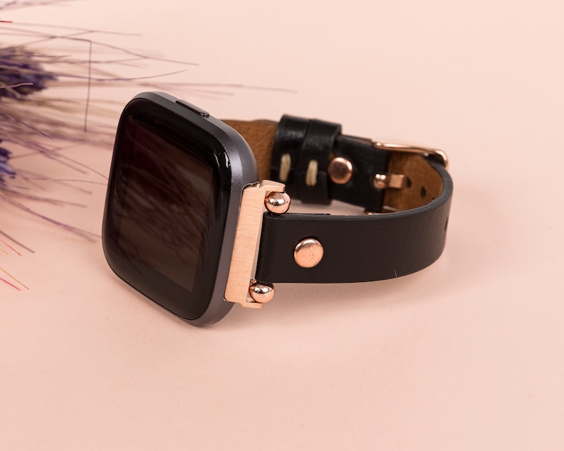 May include: A smartwatch with a dark gray square screen and a black leather strap. The watch features rose gold metal hardware, including the connector lugs, a circular stud on the strap, and a metal buckle closure for the wristband.