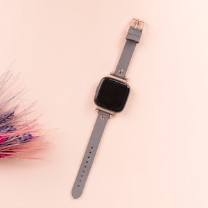 May include: A gray leather smartwatch with a black face and gold accents. The watch is on a light pink background.