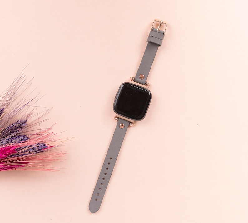 May include: A gray leather smartwatch with a black face and gold accents. The watch is on a light pink background.