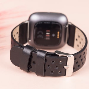 May include: Black leather watch strap with a perforated design and a silver buckle. The strap is compatible with a Fitbit Versa smartwatch.