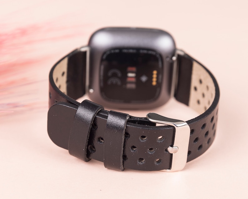 May include: Black leather watch strap with a perforated design and a silver buckle. The strap is compatible with a Fitbit Versa smartwatch.
