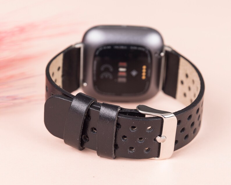 May include: Black leather watch strap with a perforated design and a silver buckle. The strap is compatible with a Fitbit Versa smartwatch.