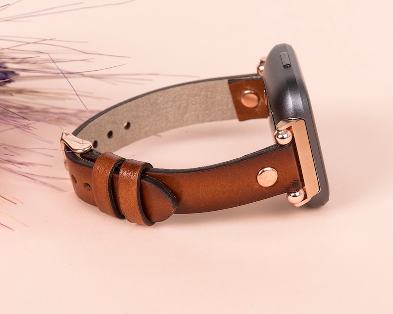 Puede incluir: Una correa de reloj de cuero marr&oacute;n con detalles en oro rosa y cierre de hebilla.
