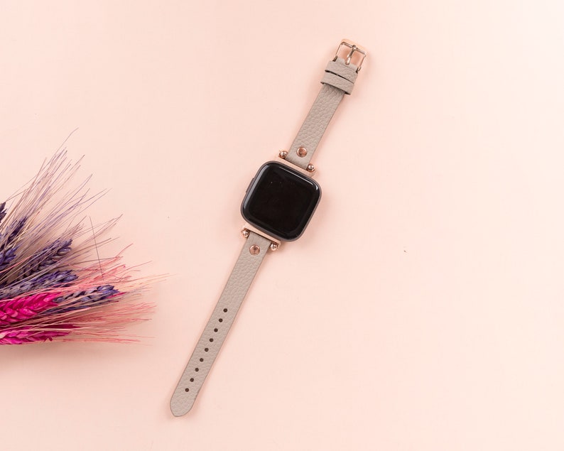 May include: A gray leather watch band with a rose gold buckle and a square black watch face.