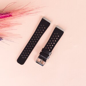 May include: Two black leather watch straps with silver buckles and a perforated design. The straps are on a light pink background.