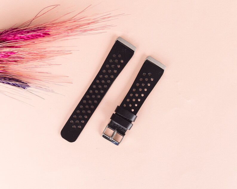 May include: Two black leather watch straps with silver buckles and a perforated design. The straps are on a light pink background.