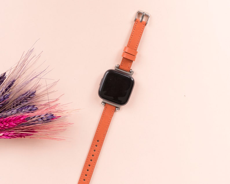 May include: A smart watch with a black face and a bright orange leather strap. The watch is on a pink background.