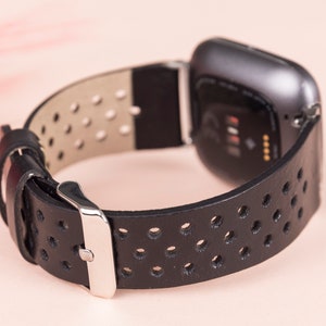 May include: Black leather watch strap with a perforated design and a silver buckle. The strap is compatible with a Fitbit Versa smartwatch.