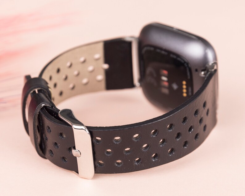 May include: Black leather watch strap with a perforated design and a silver buckle. The strap is compatible with a Fitbit Versa smartwatch.