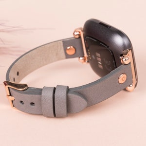 May include: A gray leather watch band with rose gold hardware. The band is designed for a Fitbit Versa smartwatch.