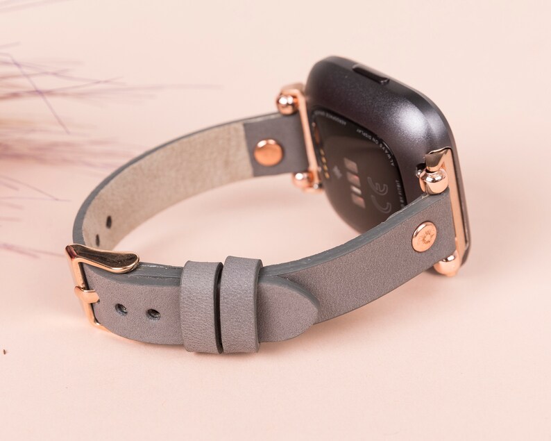 May include: A gray leather watch band with rose gold hardware. The band is designed for a Fitbit Versa smartwatch.