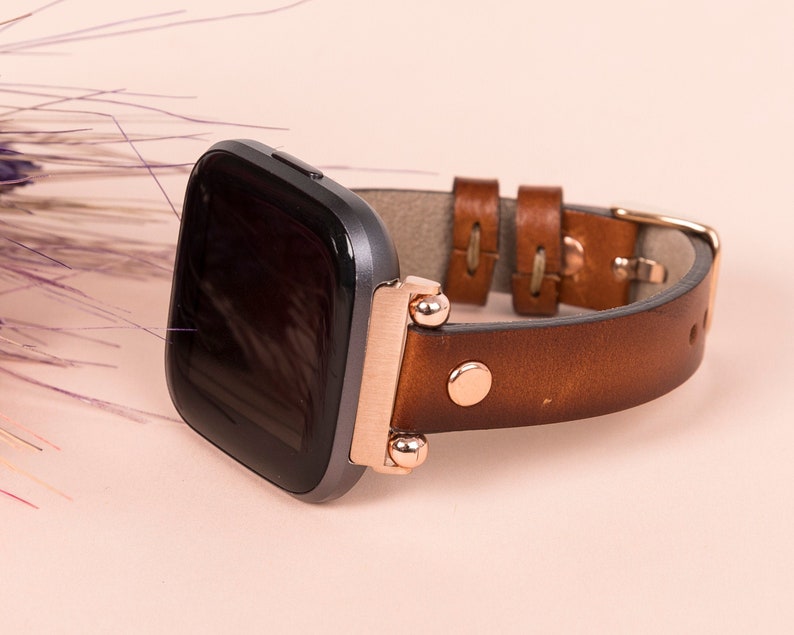 Puede incluir: Una correa de reloj de cuero marr&oacute;n con detalles en oro rosa y un reloj inteligente cuadrado negro.