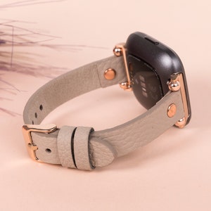 May include: A gray leather watch band with rose gold accents and a buckle closure. The band is designed for a smartwatch with a square face.