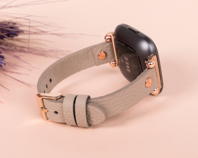 May include: A gray leather watch band with rose gold accents and a buckle closure. The band is designed for a smartwatch with a square face.