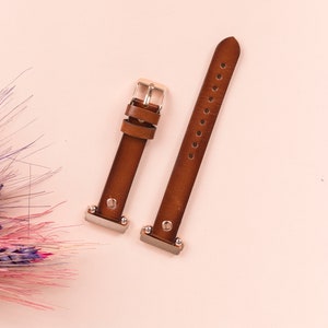 Puede incluir: Dos correas de reloj de cuero marr&oacute;n con hebillas plateadas. Las correas est&aacute;n sobre un fondo rosa claro.
