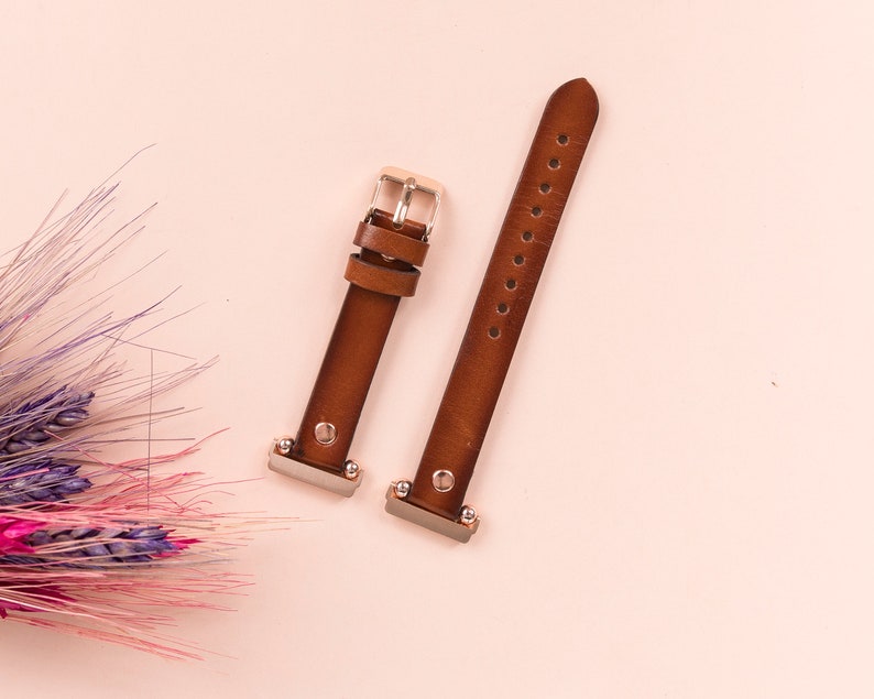 Puede incluir: Dos correas de reloj de cuero marr&oacute;n con hebillas plateadas. Las correas est&aacute;n sobre un fondo rosa claro.