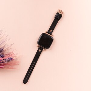 May include: A smartwatch with a black rectangular screen and a black leather strap. The watch features rose gold-toned metal hardware, including the buckle, lugs, and decorative studs connecting the strap to the watch face.