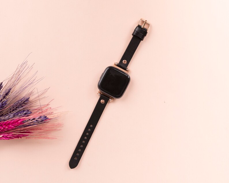 May include: A smartwatch with a black rectangular screen and a black leather strap. The watch features rose gold-toned metal hardware, including the buckle, lugs, and decorative studs connecting the strap to the watch face.