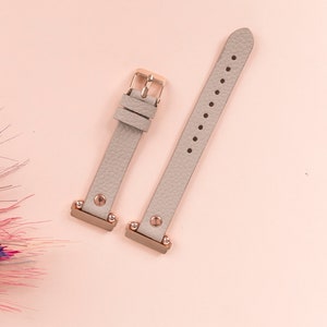 May include: Two gray leather watch bands with rose gold accents and metal clasps. The bands are on a pink background.