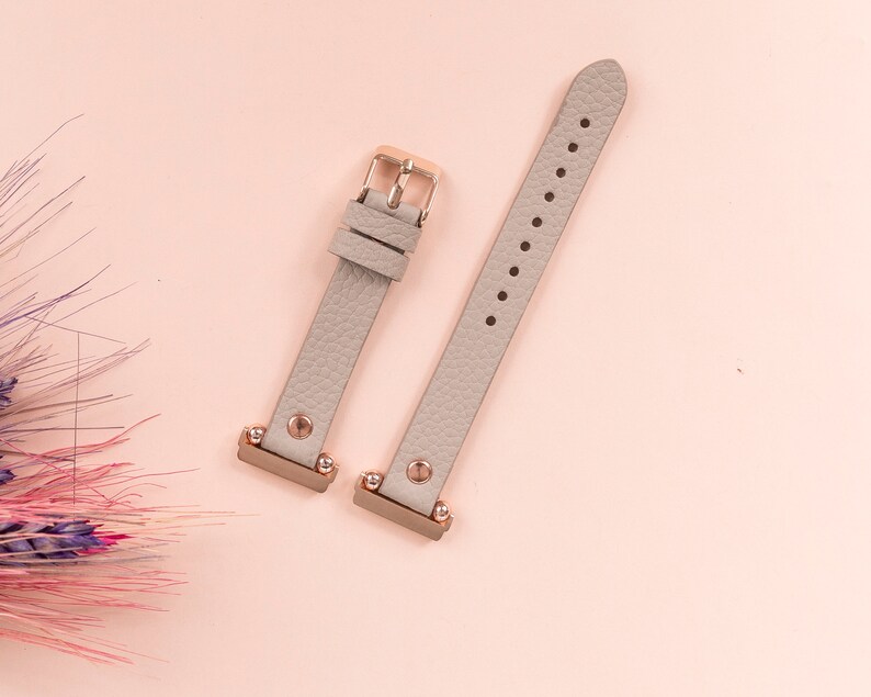 May include: Two gray leather watch bands with rose gold accents and metal clasps. The bands are on a pink background.