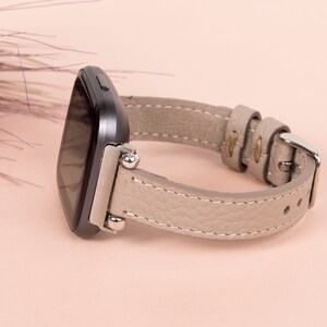 Puede incluir: Una correa de reloj de cuero gris con herrajes plateados y un reloj inteligente negro.