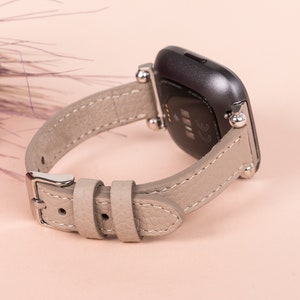 Puede incluir: Una correa de reloj de cuero gris con herrajes plateados y cierre de hebilla. La correa est&aacute; dise&ntilde;ada para adaptarse a un reloj inteligente.
