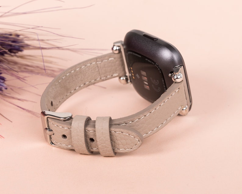 Puede incluir: Una correa de reloj de cuero gris con herrajes plateados y cierre de hebilla. La correa est&aacute; dise&ntilde;ada para adaptarse a un reloj inteligente.