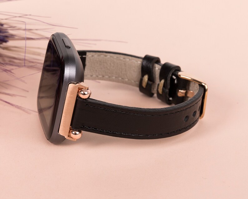 May include: A black leather watch strap with rose gold accents and a square-shaped watch face.