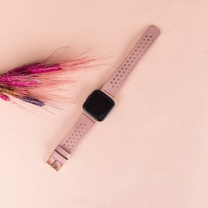 Puede incluir: Un rastreador de actividad f&iacute;sica rosa con una pantalla negra y una banda rosa perforada. El rastreador est&aacute; sobre una superficie rosa claro.